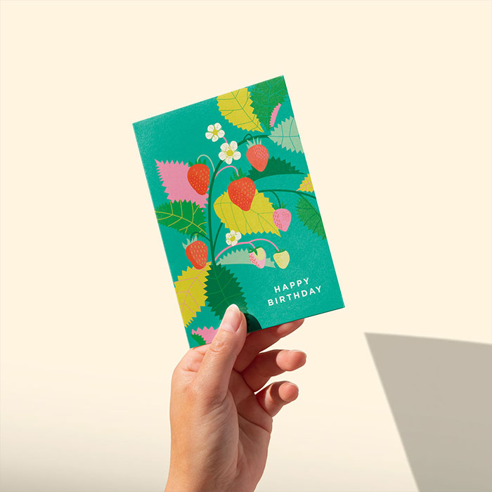 A hand holds a greetings card featuring an illustration of strawberries and the words 'happy birthday'